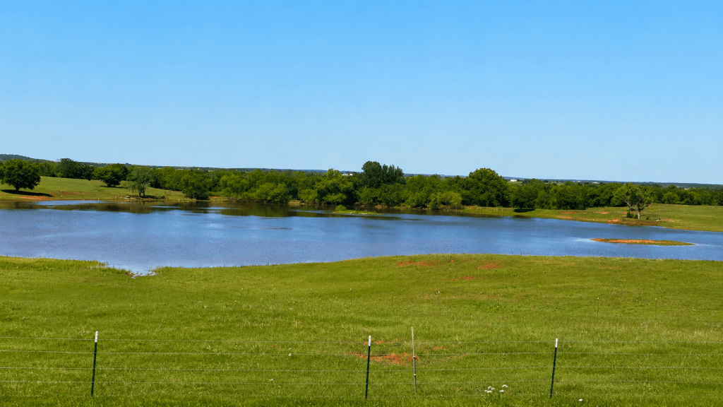 A calm pond in the middle of a green Oklahoma pasture, with trees lining the far edge.