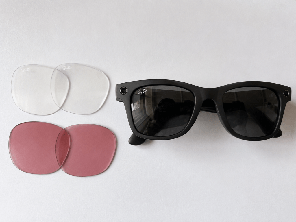 A pair of Ray-Ban Meta smart glasses with the dark lenses installed, and clear and pink replacement lenses lying beside them, ready to be swapped in.