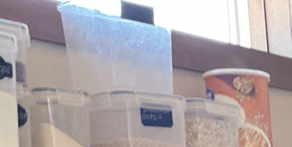 A pantry shelf holding food-storage containers of different shapes and sizes — several clear plastic tubs and a round canister — kept intentionally different so they can be told apart by feel.